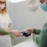 Pacjent płaci kartą przy recepcji medycznej, obie osoby w maseczkach